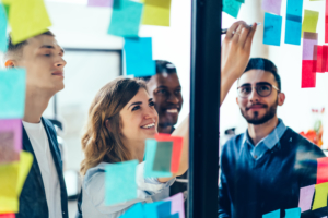 Diverse team of positive young people laughing while working together during brainstorming and standing behind glass wall with sticky colorful papers.Cheerful students learning words from stickers